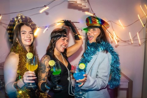 Three diverse women wearing festive costumes, decorations, and holding colorful cocktails, enjoying