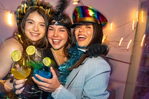Three happy young women laughing and toasting with colorful cocktails, dressed in costumes,