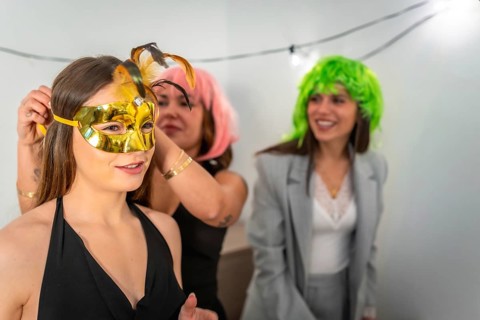 Friends preparing for a celebration, with one woman helping another put on a gold masquerade mask