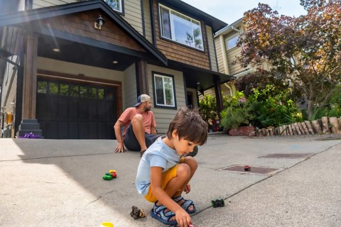 Young boy plays with toy cars on the driveway of a modern home while his father watches from