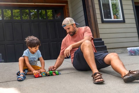 Father and son are sitting on the ground in their driveway, playing with a collection of toy cars,