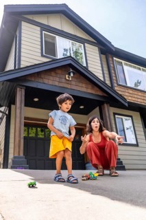 Mother and son are crouching down, playing with toy cars on the driveway in front of their house,