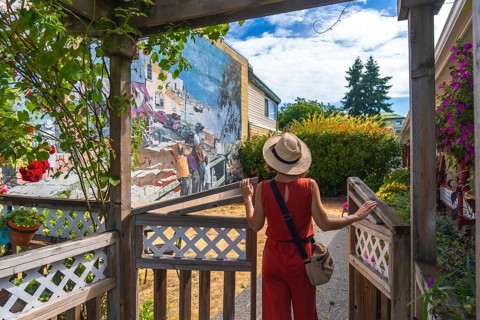 Female tourist in a straw hat and red jumpsuit opening a wooden gate to admire a vibrant mural