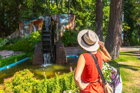 Tourist wearing a straw hat holds the brim while admiring a water wheel in a garden setting in