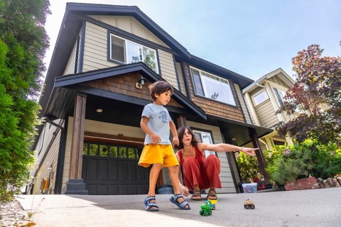 Mother crouches next to her son, pointing at toy cars on the driveway of their two story home,