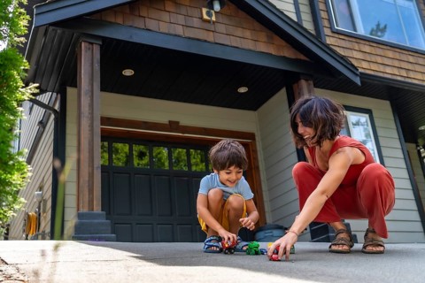 Mother and her young son are happily playing with toy cars on the driveway of their modern suburban