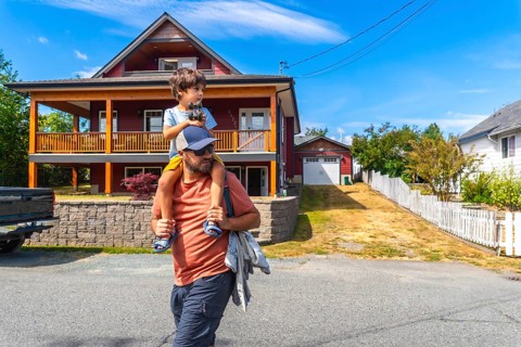 Father carrying son on shoulders while walking past a two story house with a porch and detached
