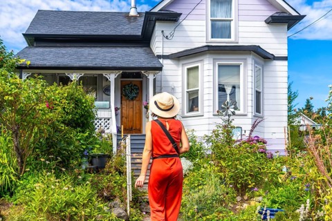 Tourist wearing a straw hat and an orange jumpsuit walks up a garden path towards a charming house