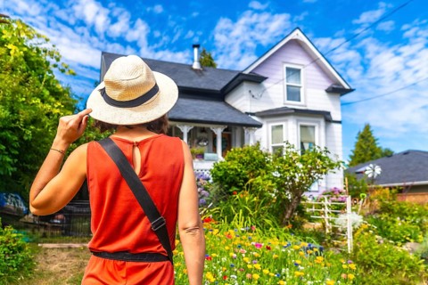 Female tourist wearing a straw hat, holding it with one hand, admiring a beautiful blooming garden