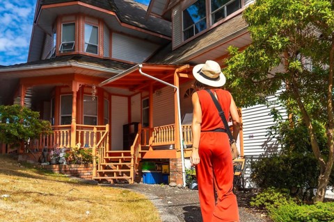 Tourist wearing a straw hat and a red jumpsuit is walking on a path near a traditional wooden house