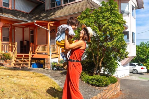 Happy mother wearing a straw hat and an orange jumpsuit lifting up her cheerful son in the yard of
