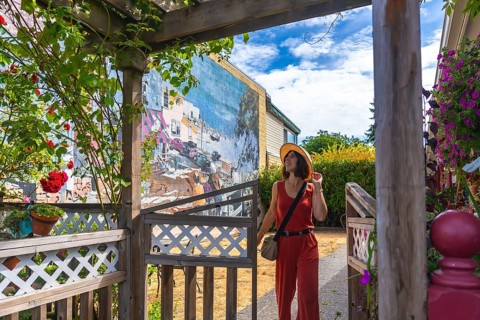 Female tourist wearing jumpsuit and straw hat walking through wooden gate and admiring colorful