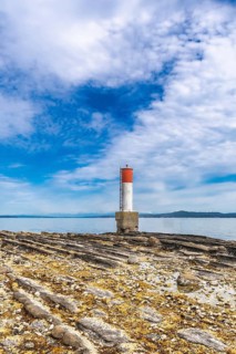 Red and white lighthouse standing on a rocky beach with a blue sky and clouds in the background in