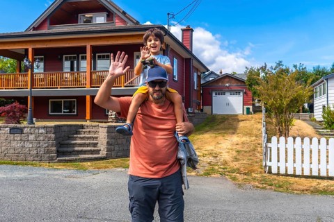 Father carrying his son on his shoulders, both waving, standing in front of their house in
