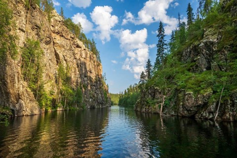 A calm lake lies in a wooded gorge, surrounded by tall rocks and reflecting the sky covered with