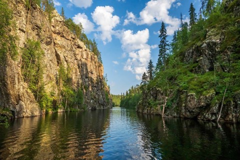 A calm lake lies in a wooded gorge, surrounded by tall rocks and reflecting the sky covered with