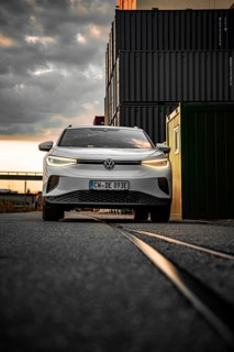 White car drives past container warehouse at sunset, dramatic urban scene, VW ID4 electric car,