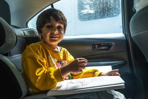 Young child happily drawing in a coloring book while secured in a car seat during a road trip,