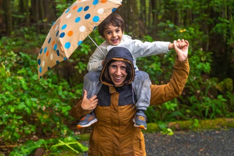 Mother carries her smiling son on her shoulders under a colorful umbrella, both wearing raincoats