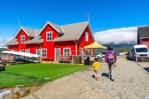 Mother and son are walking on a gravel path near a vibrant red house with white trim, canoes