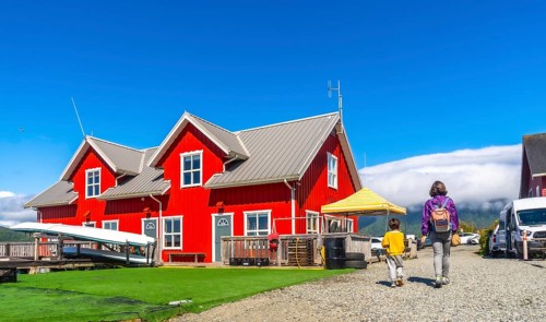 Mother and son walking on gravel path near vibrant red wooden building with canoes, enjoying summer