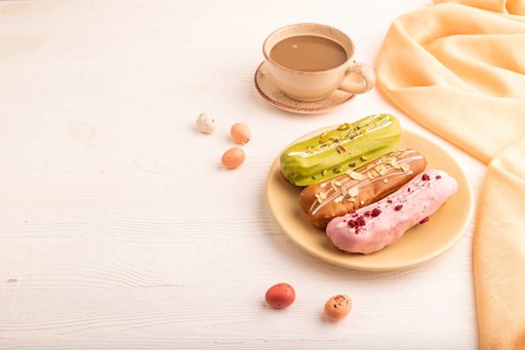 Glazed Eclairs on white wooden background and orange linen textile, cup of coffee, side view, copy