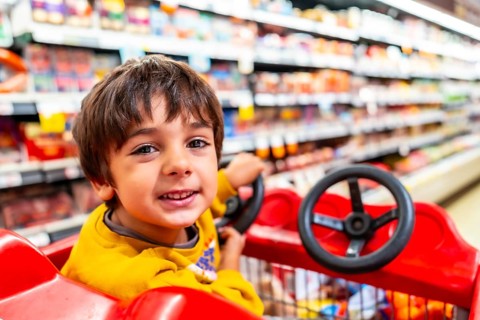 Cheerful child is enjoying a shopping trip, sitting in a shopping cart with a toy steering wheel