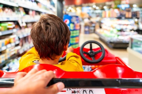 Parent pushes a shopping cart designed as a toy car, with their child pretending to drive,
