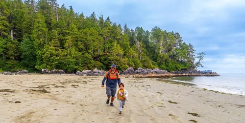 Father and son running together on a sandy beach with lush green forest and rocky coastline in