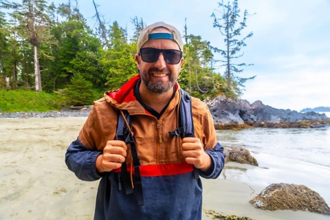 Happy tourist wearing sunglasses and baseball cap is holding his backpack straps while enjoying a
