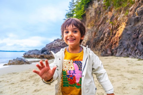 Happy child waving on a sandy beach with rocky cliffs and ocean in the background, enjoying a sunny