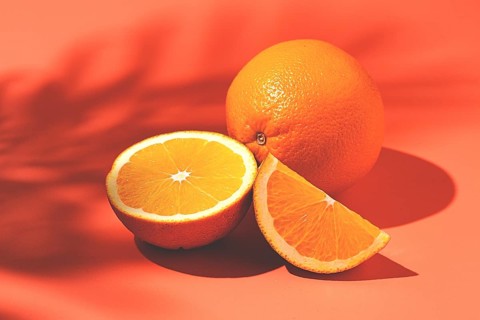On the coral surface, next to two neatly sliced slices, there is a whole orange. Sunlight casts