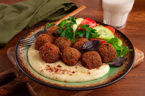 A plate holds falafel balls, tahini, salad with tomatoes and cucumbers, and a drink. The setting is