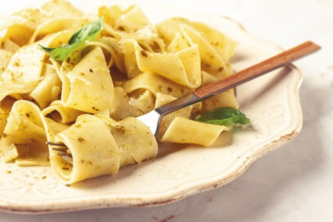 Fresh papardelle pasta, with basil and pesto sauce, is served, on a white plate, with a fork held