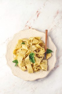 Fresh papardelle pasta, with basil and pesto sauce, is served, on a white plate, with a fork held