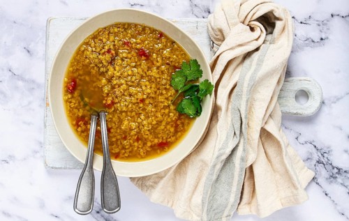 A bowl of delicious lentil soup sits on a marble countertop, garnished with fresh herbs. Silver