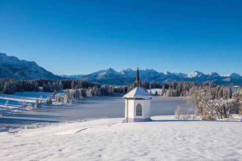 Snowy landscape with chapel and mountains in the background under clear blue sky, Hegratsrieder