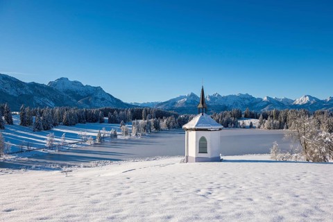 Small chapel in front of snow-covered mountains and forests under a clear sky, Hegratsrieder See,