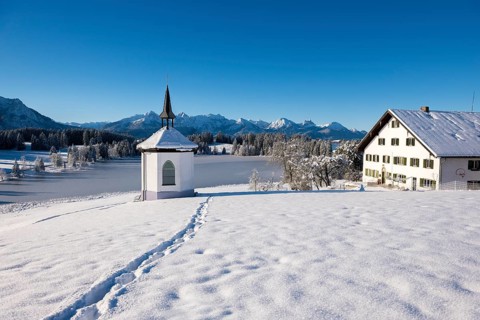 Snowy landscape with chapel and farmhouse against mountain backdrop under blue sky, Hegratsrieder
