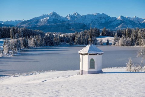 Chapel on the shore of a frozen lake surrounded by snow and mountains under a blue sky,