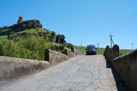 A car drives over a stone bridge with a building on a green hill in the background under clear sky,