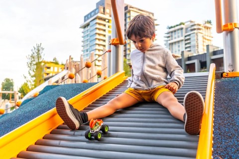 Young boy sitting on a playground slide, gazing at his toy monster truck while enjoying a sunny day