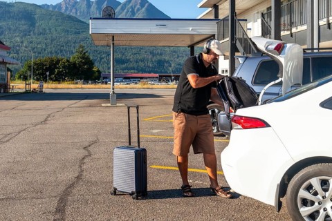 Tourist wearing headphones is loading luggage into car trunk in motel parking lot, preparing for
