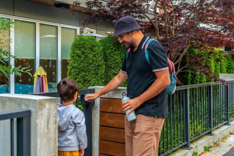 Father holding a reusable water bottle and wearing a bucket hat opens the gate for his son at a