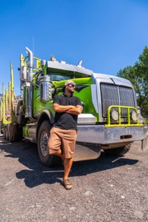 Truck driver confidently posing with arms crossed, leaning against a modern green logging truck in