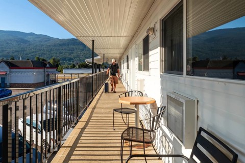 Tourist walking along a motel balcony, rolling luggage in tow, passing by a table and chairs,
