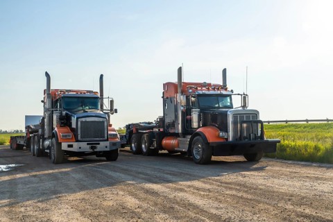 Two powerful semi trucks, gleaming in orange and black under a clear sky, parked on a dirt road