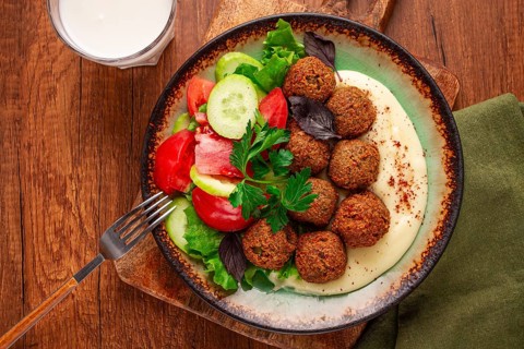 A plate holds falafel balls, tahini, salad with tomatoes and cucumbers, and a drink. The setting is