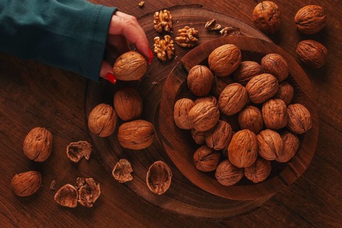 A woman's hand taking a walnut from a wooden bowl filled with walnuts. There are shells and walnuts