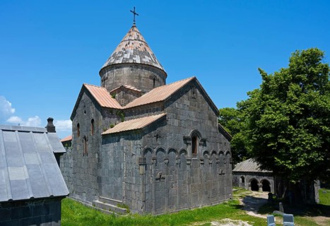 Medieval stone church with dome and cross under blue sky next to green trees, Sanahin monastery,